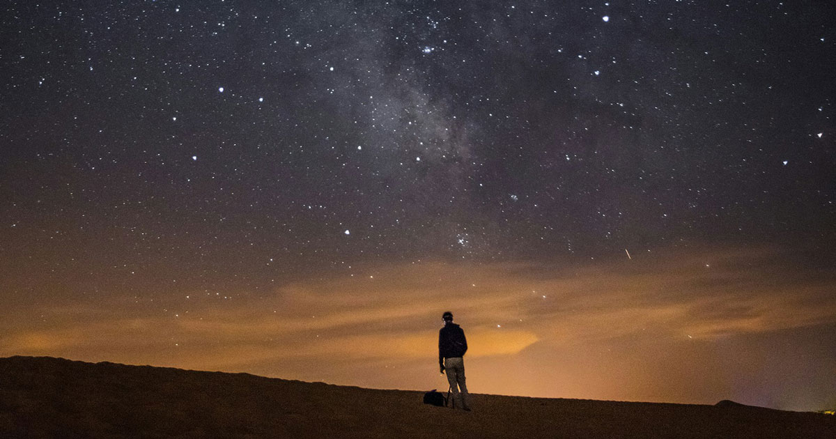 A man stands atop a sand dune under a starry night sky, silhouetted against the moonlight representing dark mode UX.